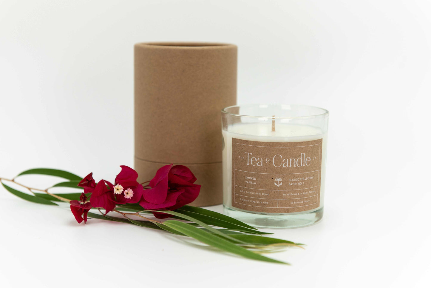 Smoked Vanilla Coconut wax candle in glass jar with luxury fragrance notes next to brown cylindrical packaging and red flowers.